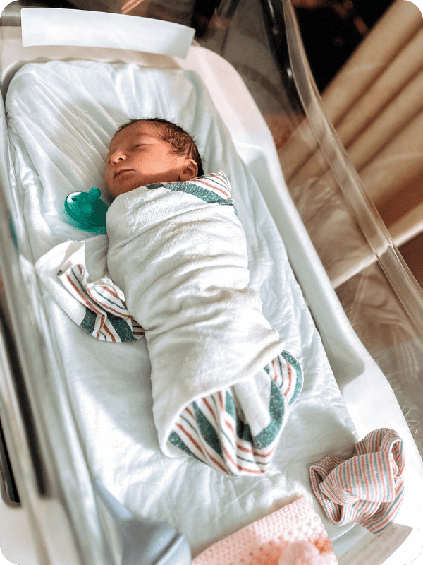 Swaddled newborn sleeps in a hospital bassinet with a pacifier laying beside them.