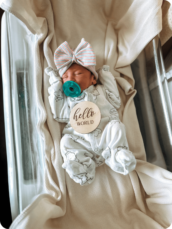 Newborn infant sucks on a pacifier and wears a bow and onesie in their hospital bassinet. There is a sign on baby's tummy that says "hello world."