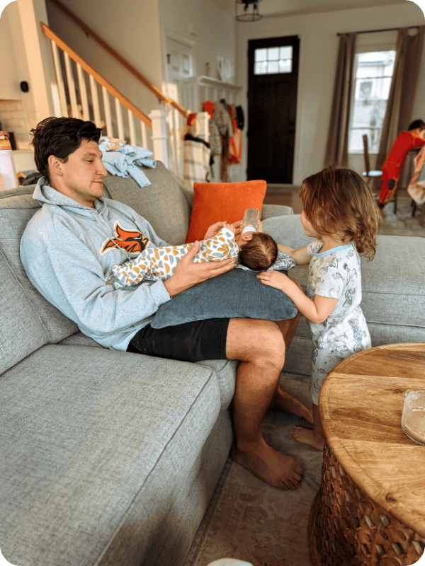 Dad sits on the couch to bottle feed newborn while big sister stands nearby to help.
