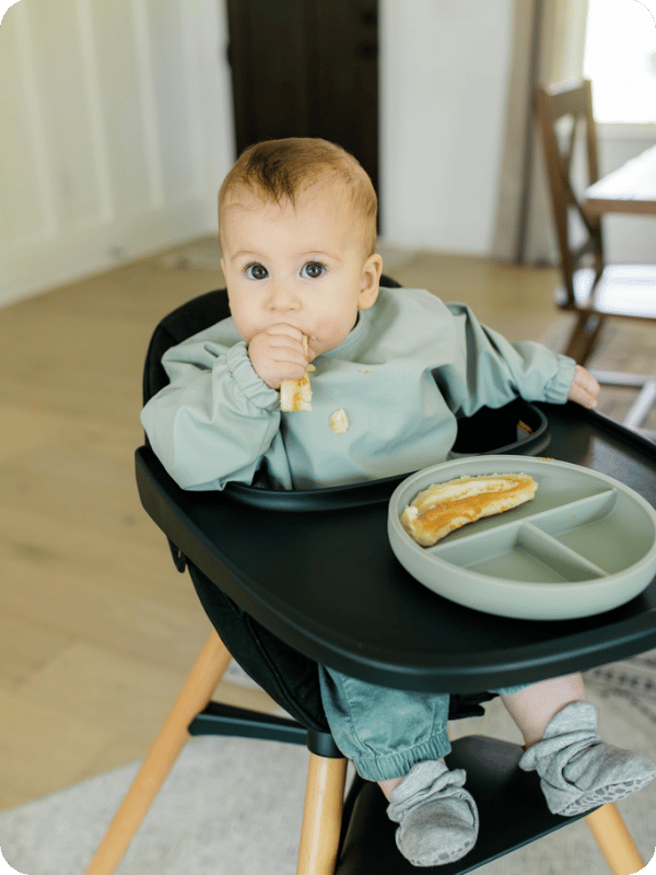 Baby looks at the camera while eating a slice of banana in the high chair.