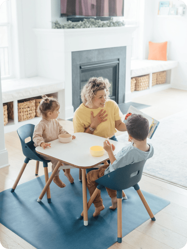 Mom reads a book to her toddlers at a kid-sized table while they eat a snack.
