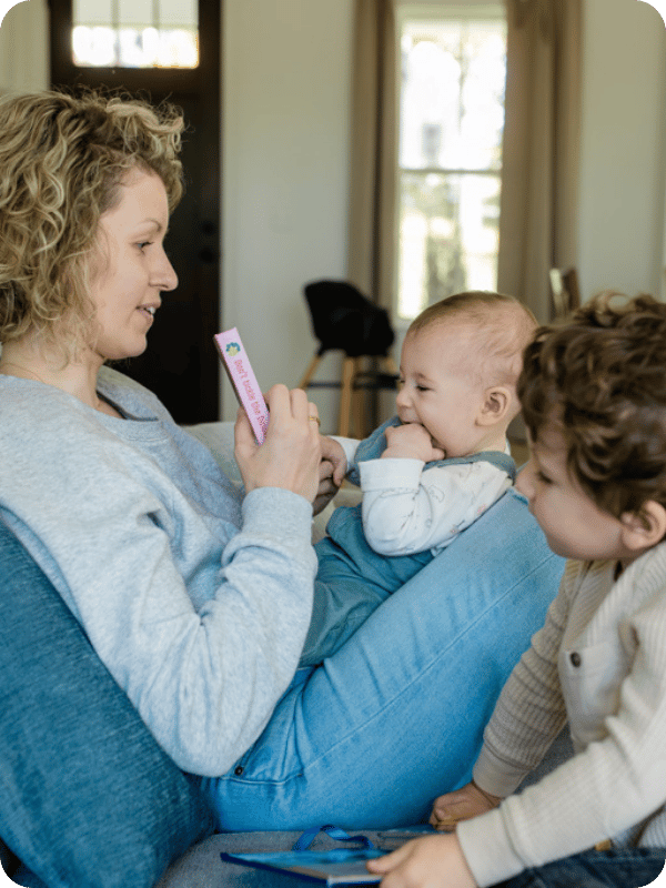 Infant sits in mom's lap facing her while mom holds up a book. Older brother sits beside them. Baby is chewing on her hand.