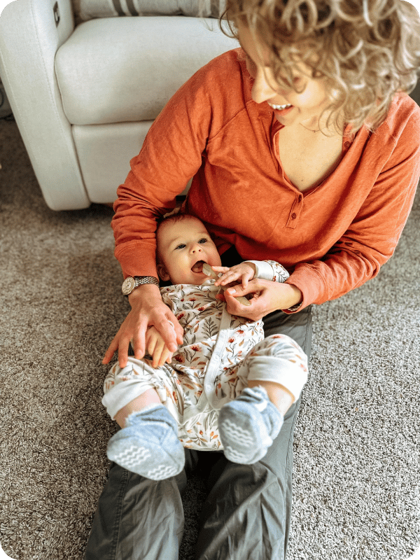 Mom brushes infant's teeth by laying her gently backward in her lap so she can see inside baby's mouth while brushing.