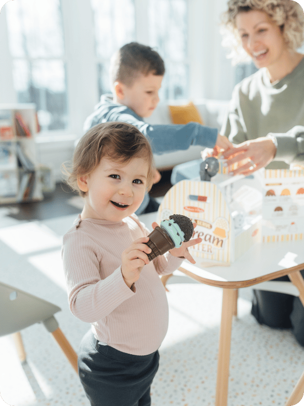 Toddler holds up a pretend play ice cream cone and smiles at the camera. Mom and older brother are smiling and playing in the background.