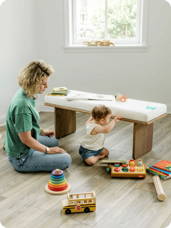 Mom sits on the floor behind infant who is reaching up to grab onto a bench to stand and play