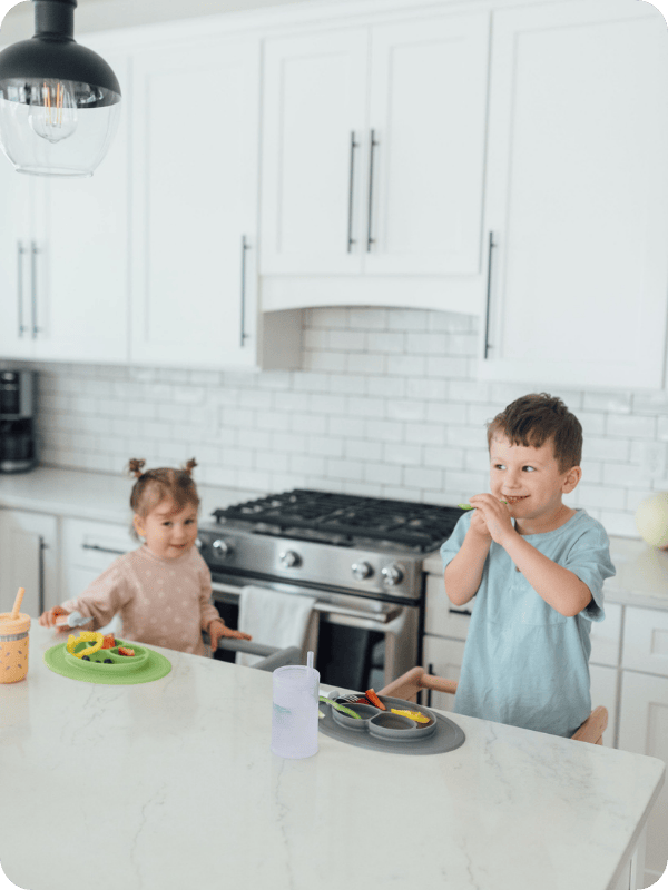 Toddlers smile at the kitchen island, standing in toddler towers to eat fruits and vegetables.