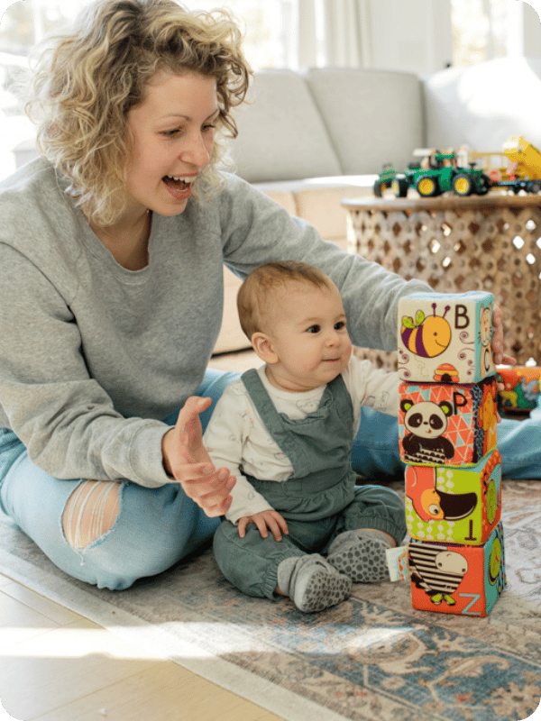 Mom sits behind baby for support as they play with blocks on the floor.