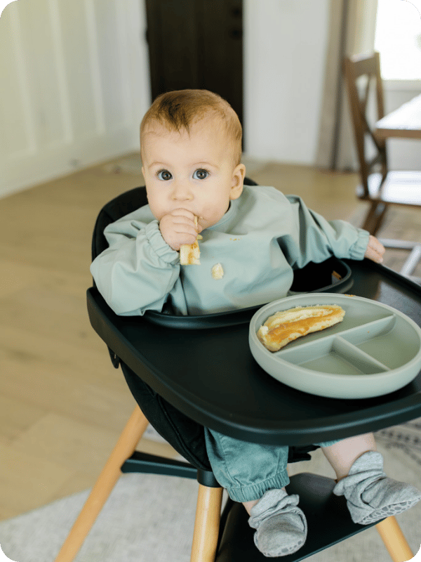 Baby eats a banana with peanut butter in her high chair.