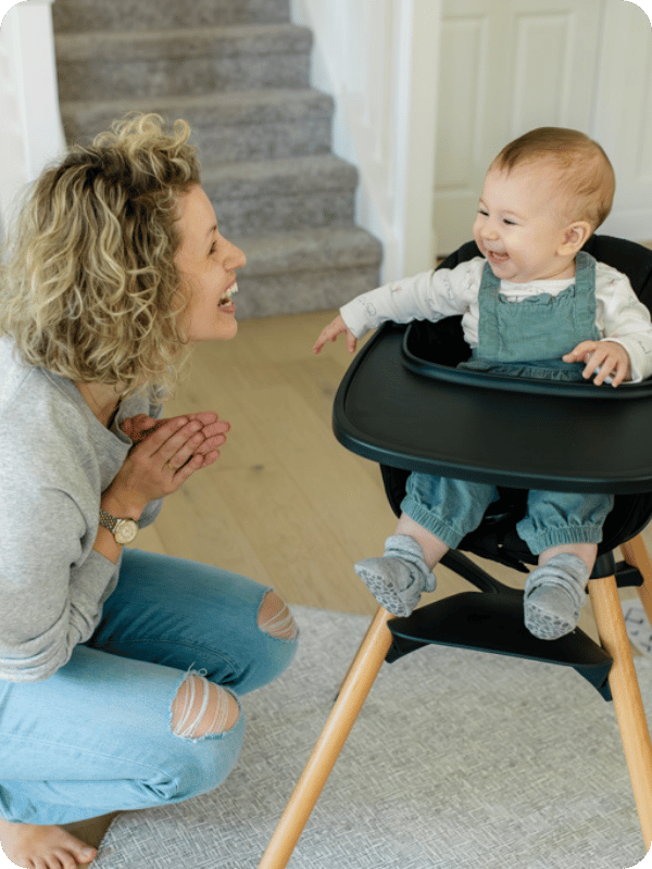 Mom and baby smile happily at each other as mom crouches to baby's eye level in the high chair before a meal.