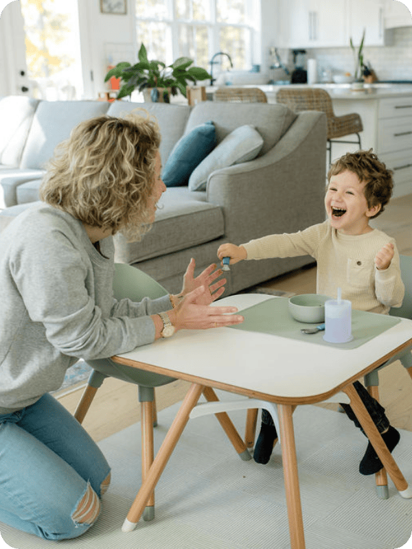 Mom and toddler laugh together while child sits at a children's table and mom is on the floor beside them