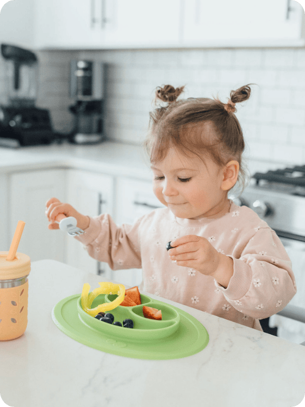 Toddler standing in a toddler tower in the kitchen, eating from a plate full of fruits and veggies