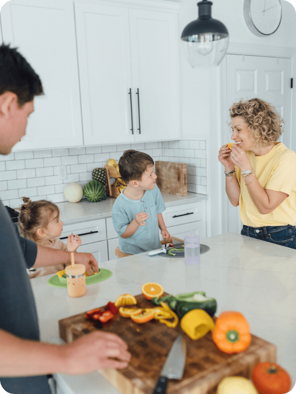 Toddlers eat orange slices with their mom in the kitchen while dad chops fruits and veggies
