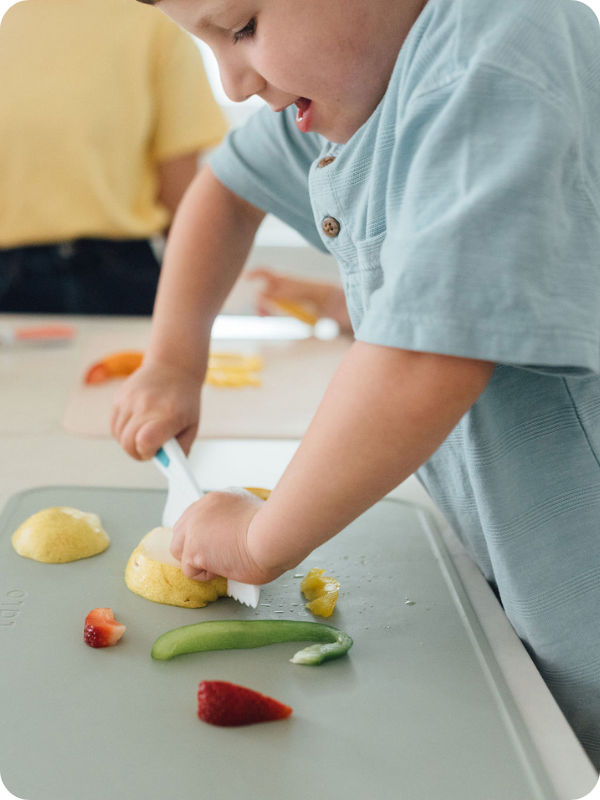 Toddler practices cutting vegetables and fruit with a child-safe knife.