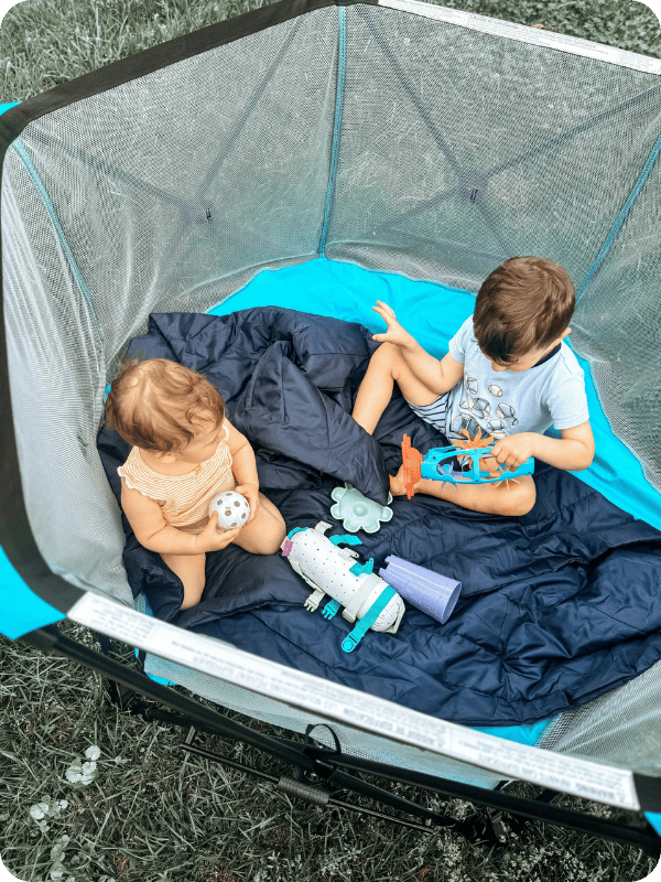 Toddler and infant play together in a playpen outside.