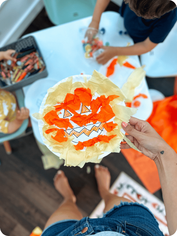 In the background, toddlers are working on crafts. A completed craft is held up toward the camera in the foreground. It is a vibrant, orange and yellow jack o lantern craft on a paper plate.