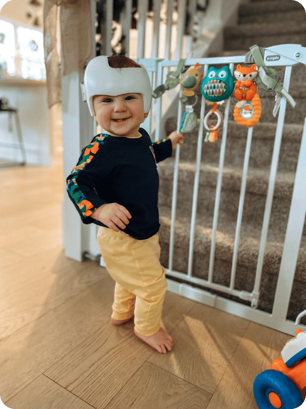 Infant stands at baby gate to play with toys. He is smiling happily at the camera and is wearing a cranial orthotic helmet.