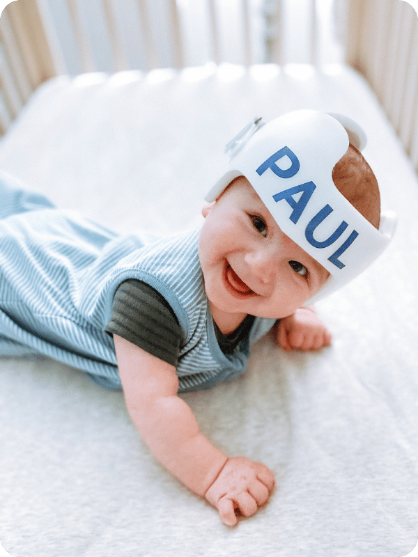 Infant wearing a sleep sack and cranial orthotic smiles up at the camera while in their crib.