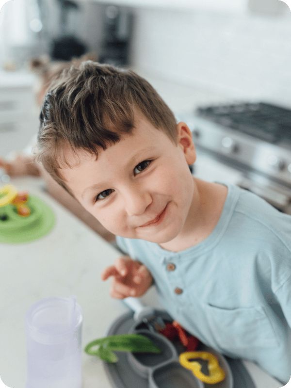 Toddler smiles at the camera while standing at the kitchen counter on his toddler tower, with a plate of snacks in front of him.