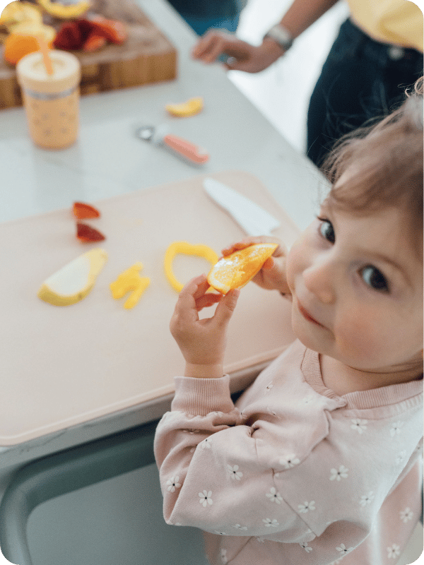 Toddler stands at the kitchen counter in her toddler tower, holding an orange slice.