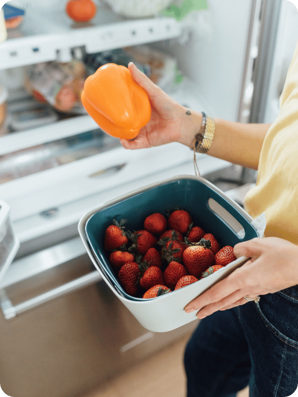 Mom stands in front of the fridge holding an orange bell pepper and carton of strawberries.