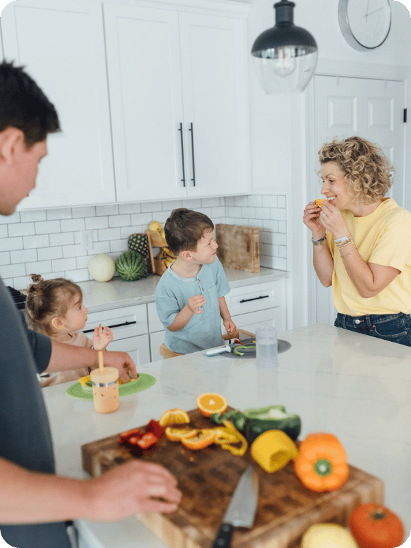 Mom and toddlers play with orange slices at the kitchen island while dad cuts veggies and fruits.