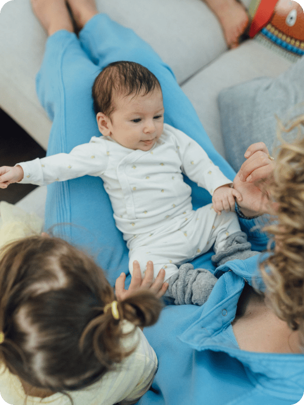 Mom holds baby on her lap while big sister sits beside them, her hand resting on baby's leg.