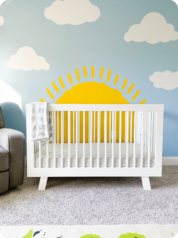 Crib against a wall with a mural of the sun and clouds.