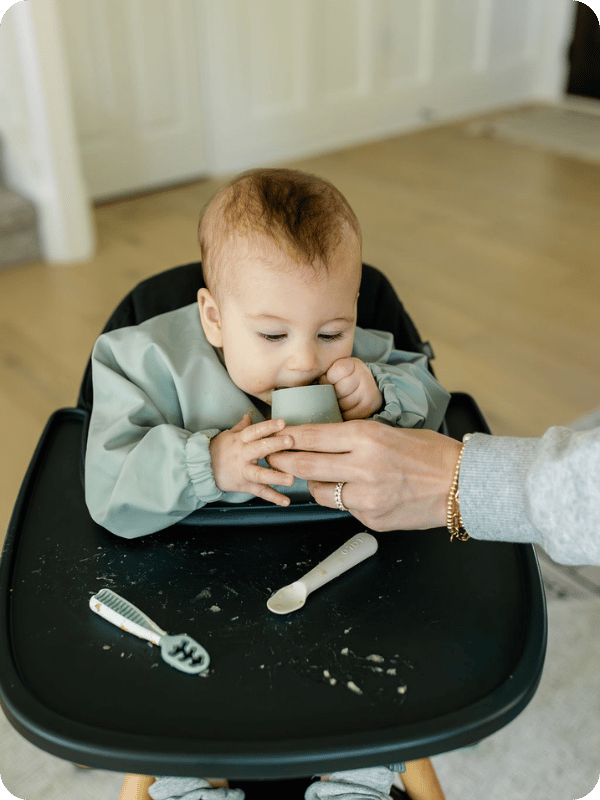 Baby practices using an open cup in the high chair, with mom's help.