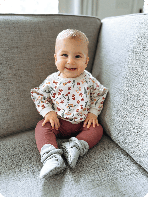 Infant sits on the couch, smiling at the camera.