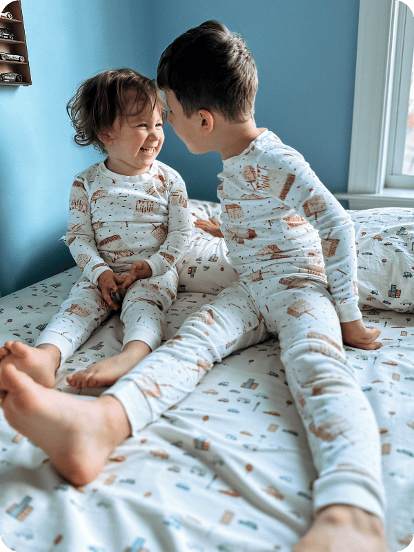 Toddlers in matching pajamas laugh together while sitting on a floor bed.