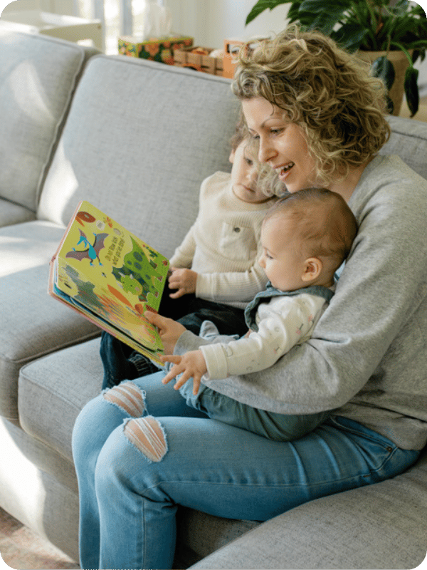 Mom reads a book with her infant and toddler while sitting together on the couch.