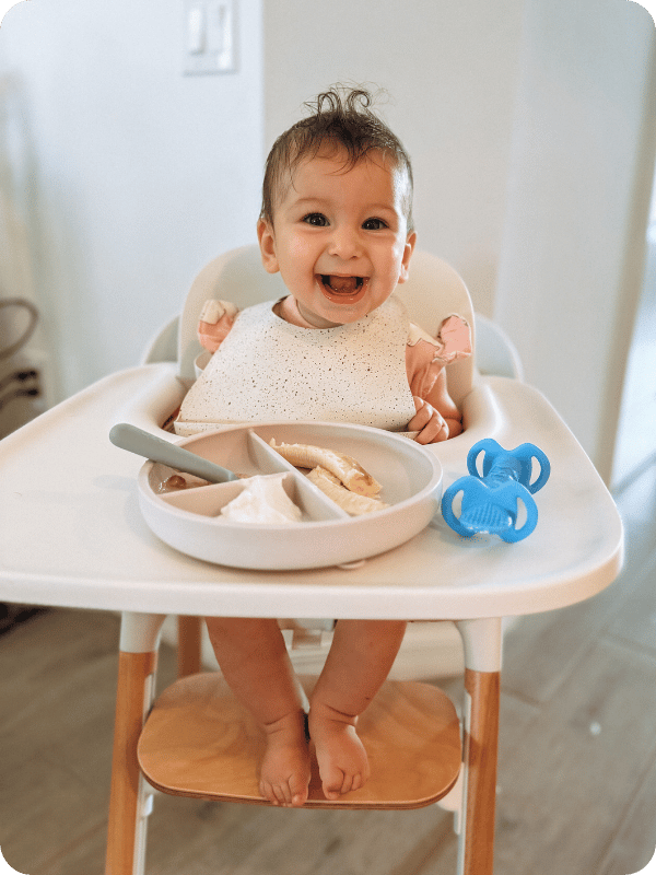 6 month old in the high chair enjoying first bites of banana. She's smiling at the camera