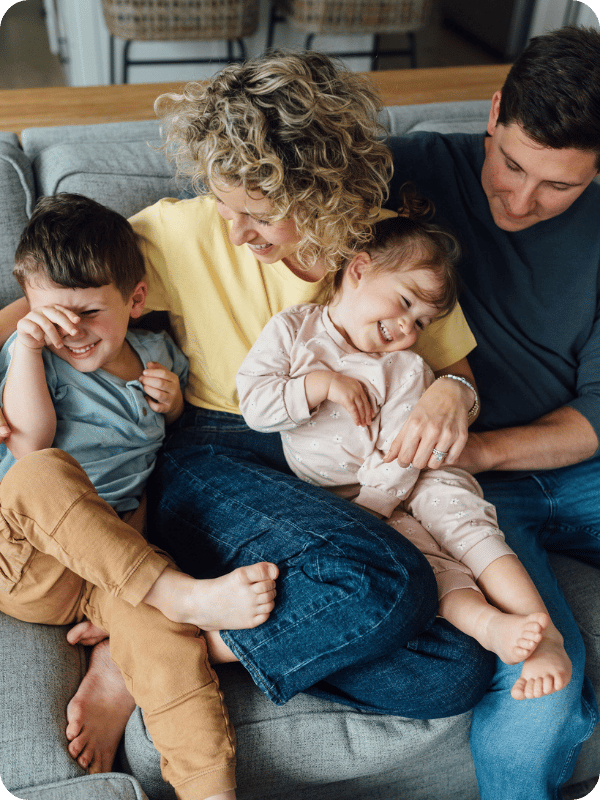 A family with mom, dad, and two toddlers laugh while sitting together on the couch.