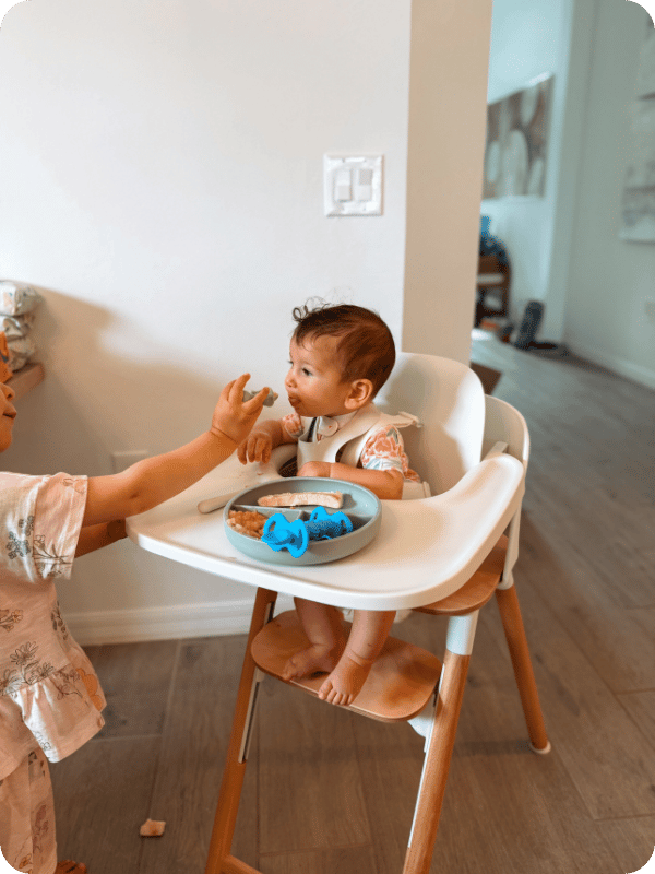 Toddler helps feed baby a meal in the high chair