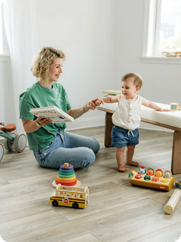 Mom sits on the floor beside young toddler, holding her hand as she looks at a book