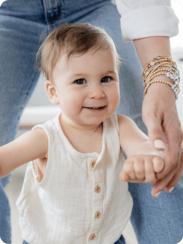Toddler smiles at the camera while mom holds her hand to walk